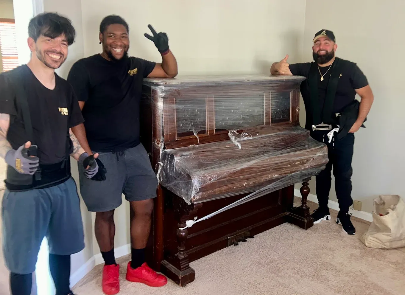 Three Premiere Movers crew members beside an upright piano wrapped in blankets and shrink wrap