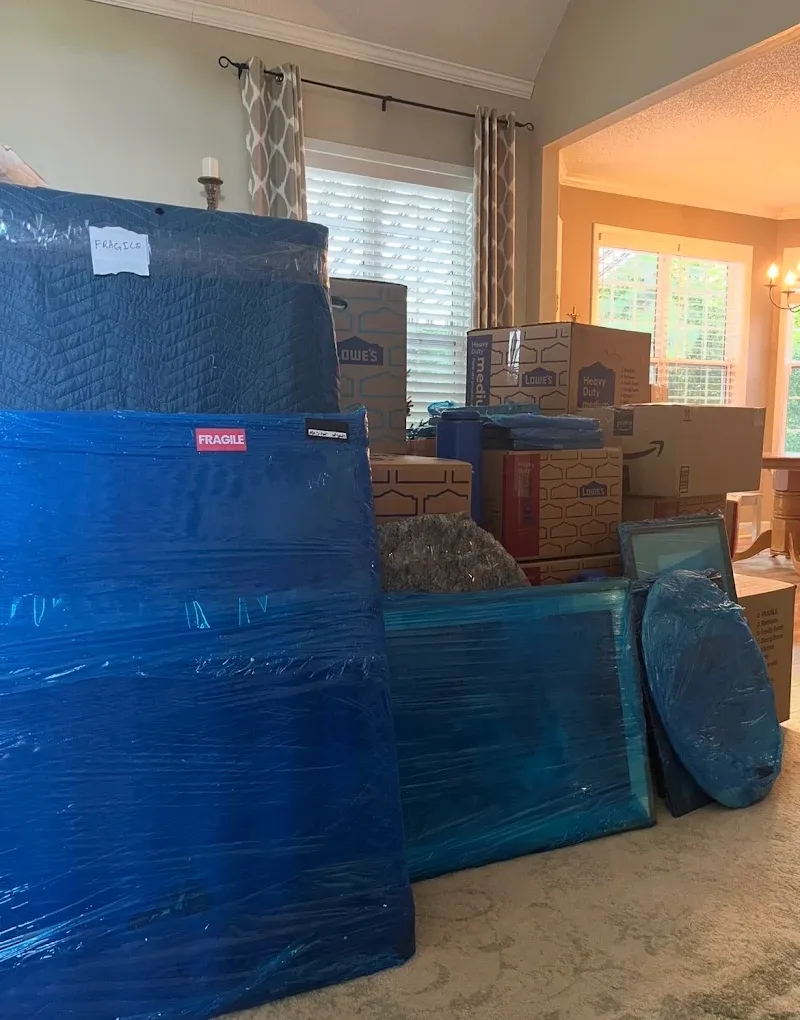 Furniture wrapped in moving blankets and shrink wrap with fragile labels, stacked beside packed boxes in a living room
