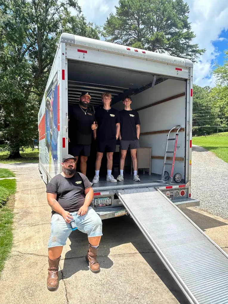 Four Premiere Movers crew members at the open back of a moving truck with the ramp down and a hand truck loaded